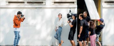 Image of a group of young people making a film, with one person taking a photo of the film crew, one holding the camera and another holding up a light reflector.