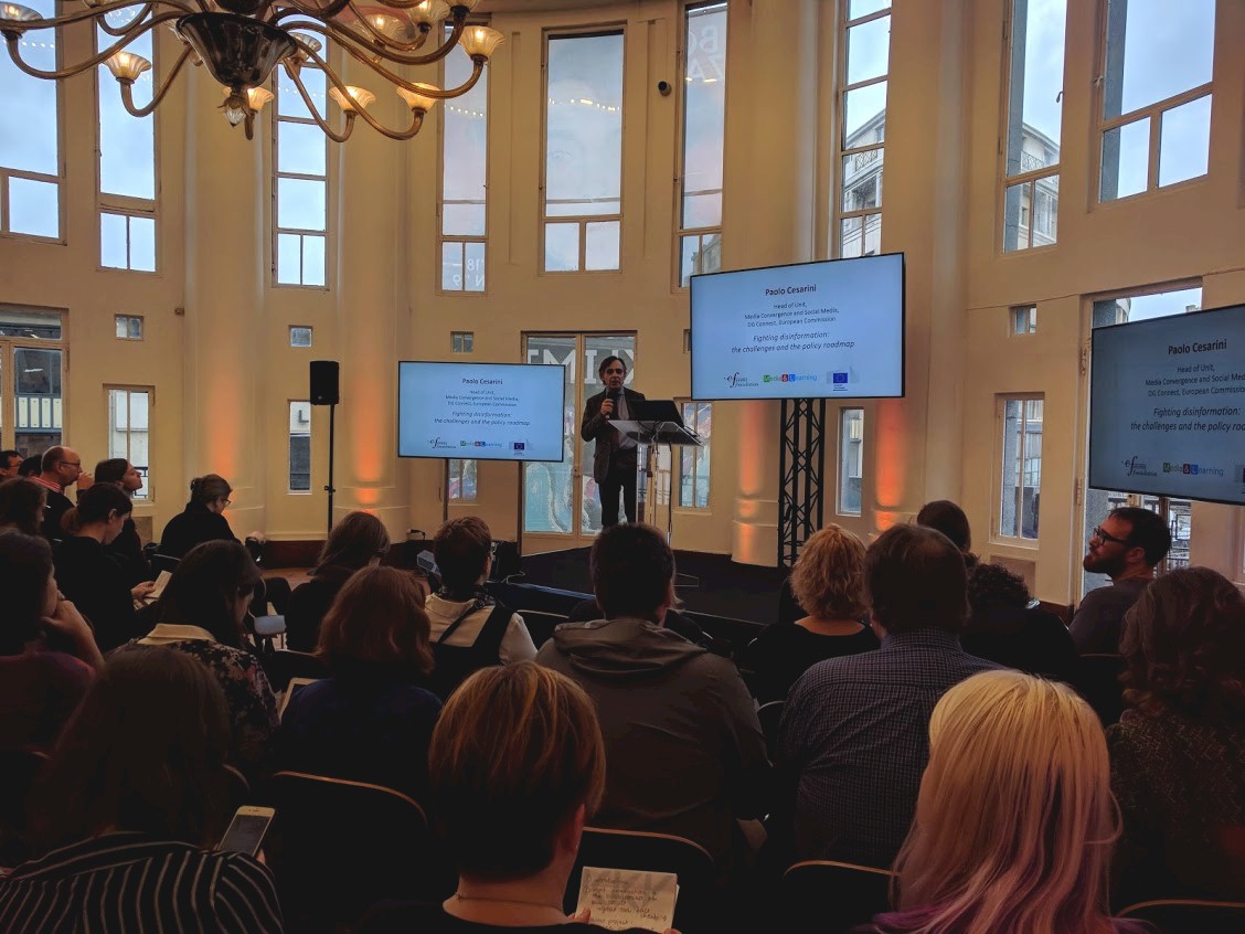 A speaker on a stage presenting to an audience, with two large screens displaying his presentation.