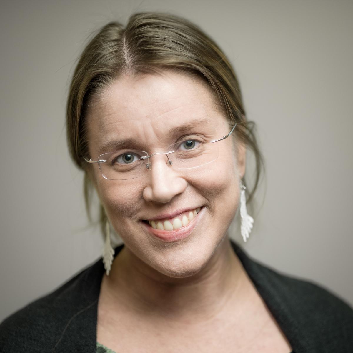Maarit Jaakkola smiling, wearing glasses and a dark top, with light brown hair parted in the middle.