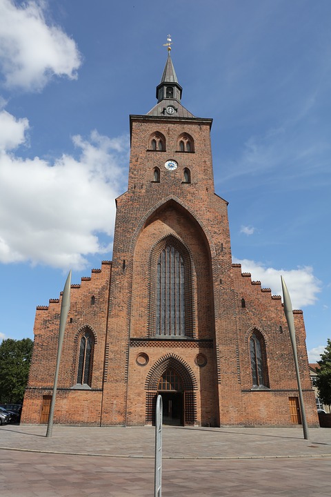 The red brick facade of Odense Cathedral, with a tall spire and arched windows, under a blue sky.
