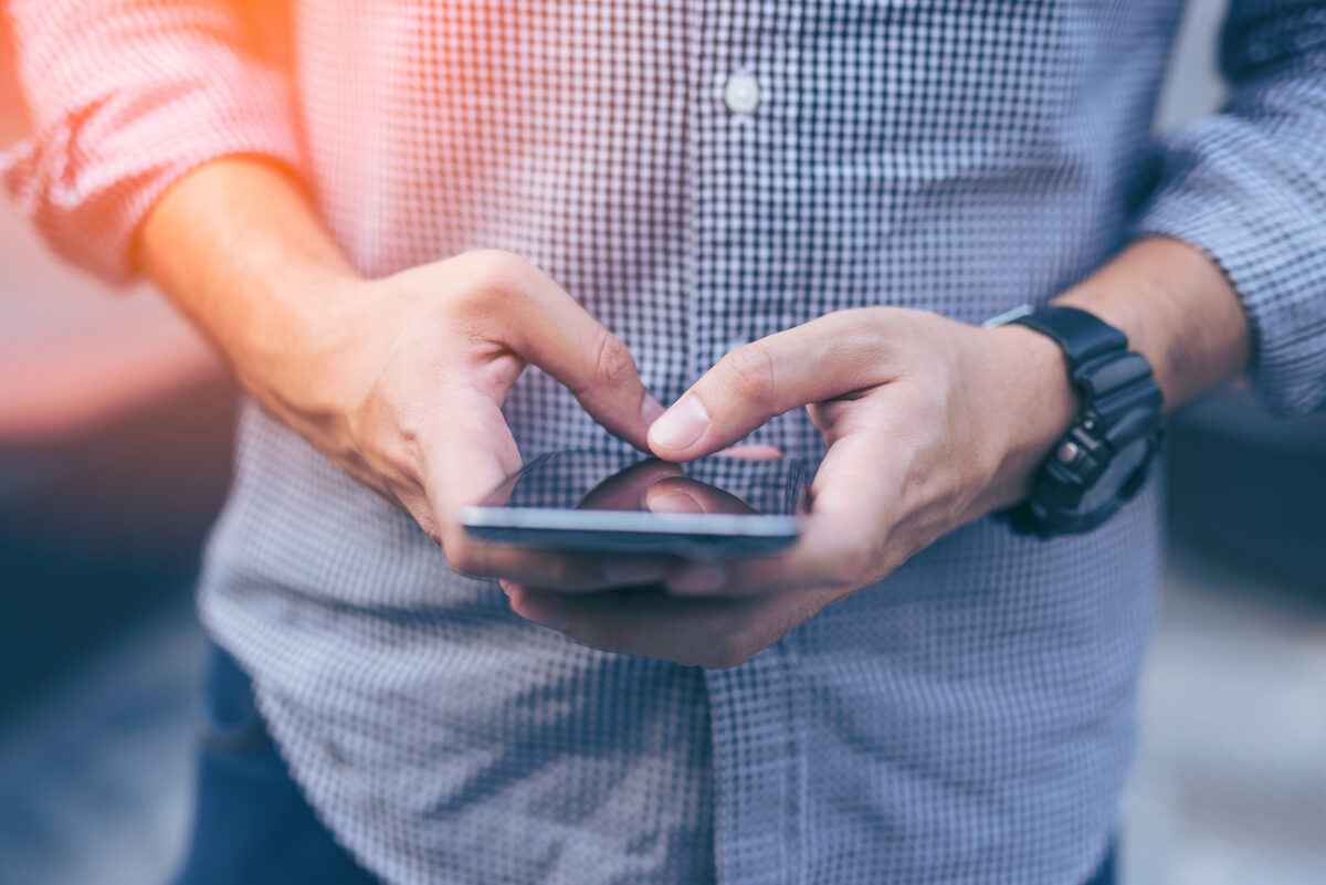 Close-up of a person's hands holding and typing on a smartphone, wearing a checkered shirt.
