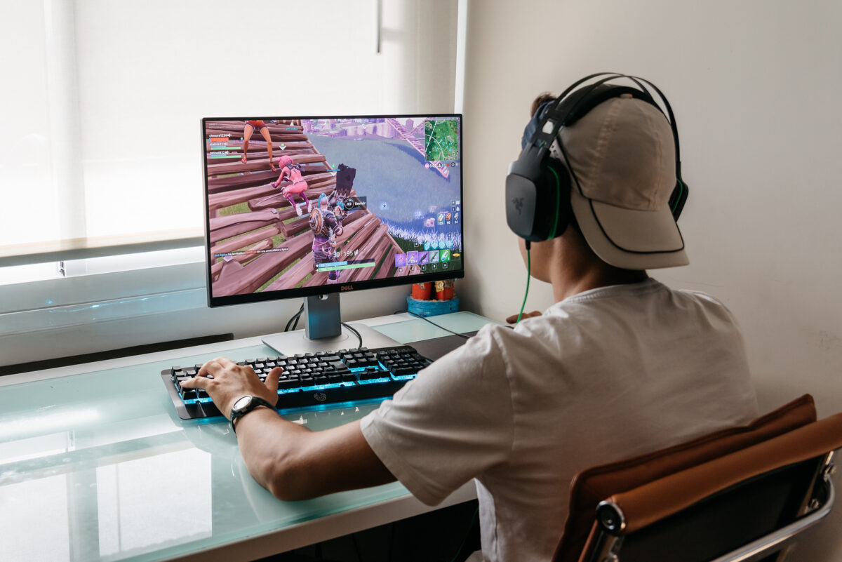 A person wearing a cap and headphones plays a video game on a computer, viewed from behind.