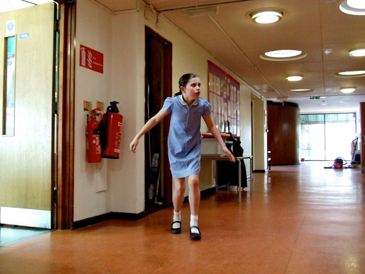 A young girl in a blue and white checked dress walks down a school corridor with arms outstretched.