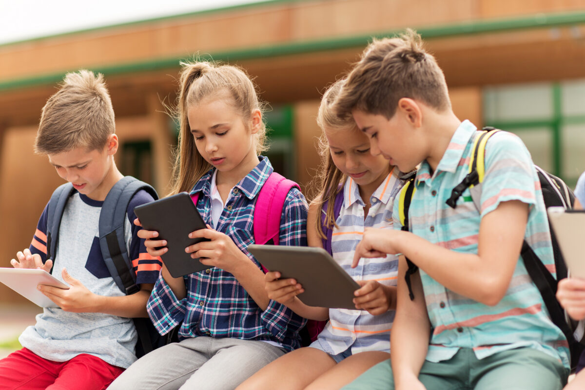 Four primary school children with backpacks sitting outdoors, looking at and interacting with tablets.