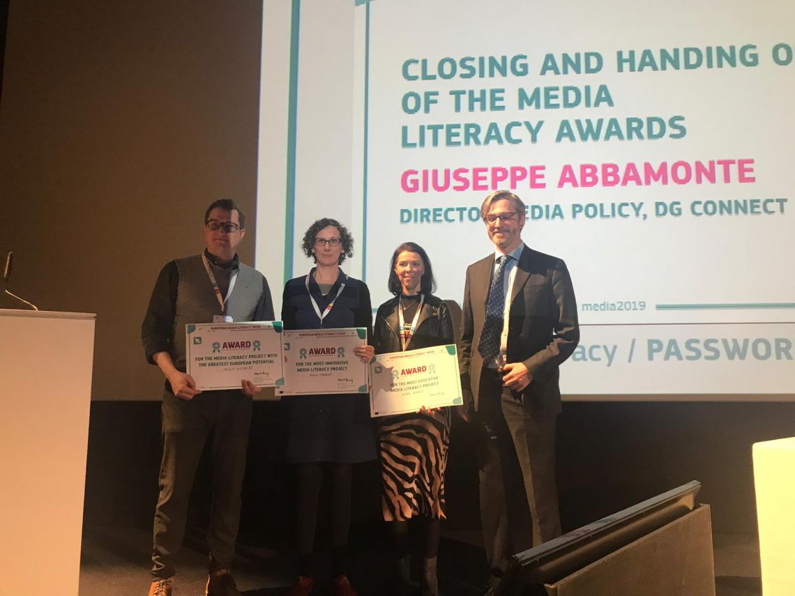 Four people on stage holding awards, with a screen behind them displaying text about media literacy awards.