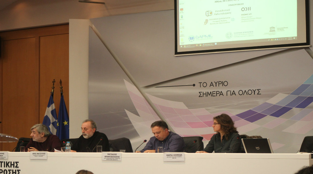 Four speakers seated at a table on a stage, with a projection screen and Greek and EU flags behind them.