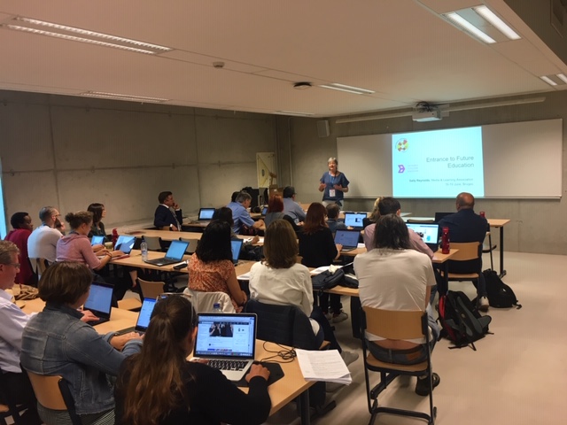 A speaker presents to a classroom of people seated at desks with laptops, a presentation slide is projected