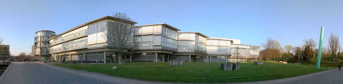 Panoramic view of a modern university building with glass facades and a round tower, surrounded by green lawns and trees.