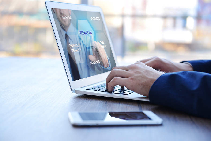 A person typing on a laptop, with a smartphone next to it. The laptop screen shows a webinar.