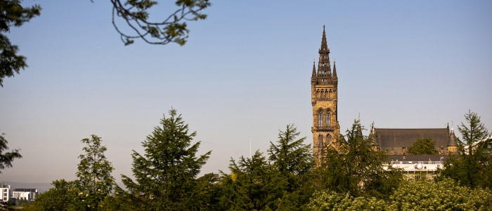 The spire of the University of Glasgow's main building rises above a canopy of green trees.