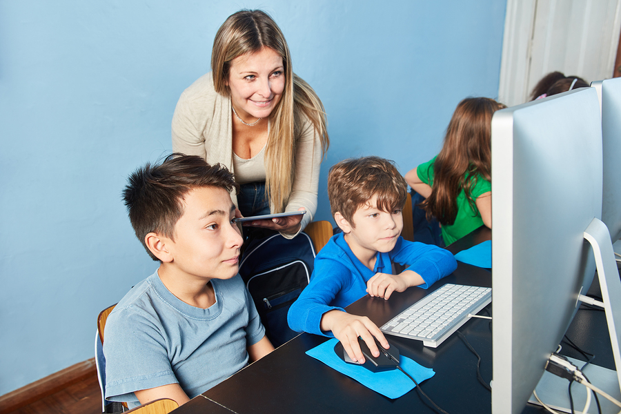 A teacher with a tablet stands behind two boys using computers in a classroom, with other children in the background.