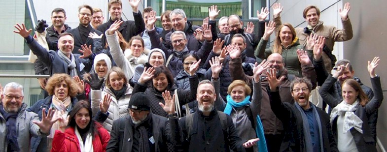 A large group of people, mostly adults, smiling and waving at the camera, some wearing coats and scarves.