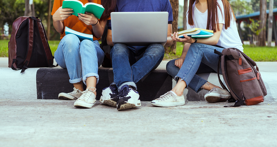 Three students sitting on a low wall, one reading a book, one using a laptop, and one holding books.