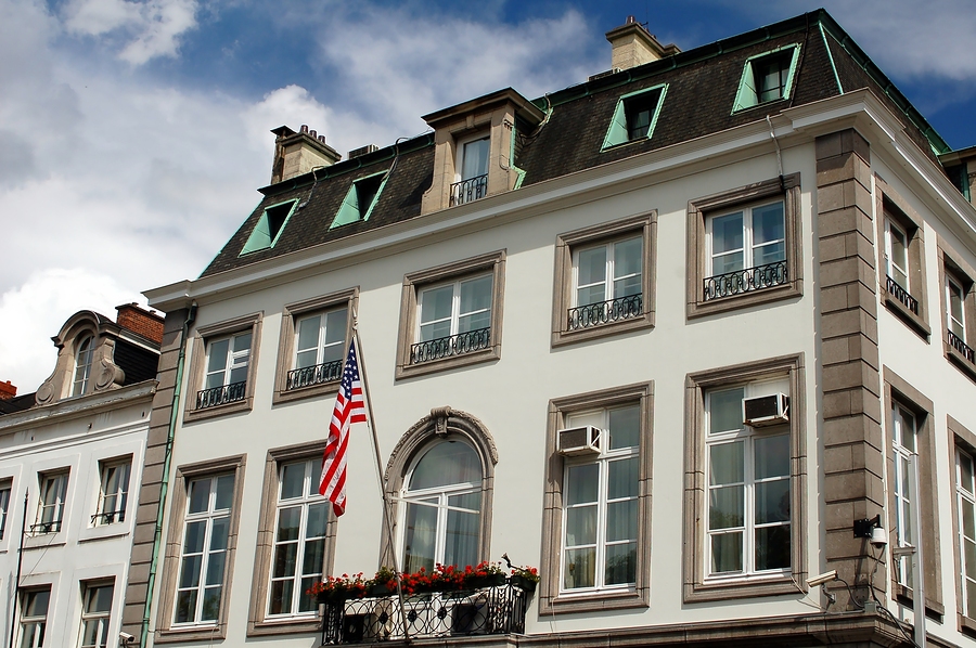 The US flag hangs from a white building with a dark roof and many windows, under a blue sky.