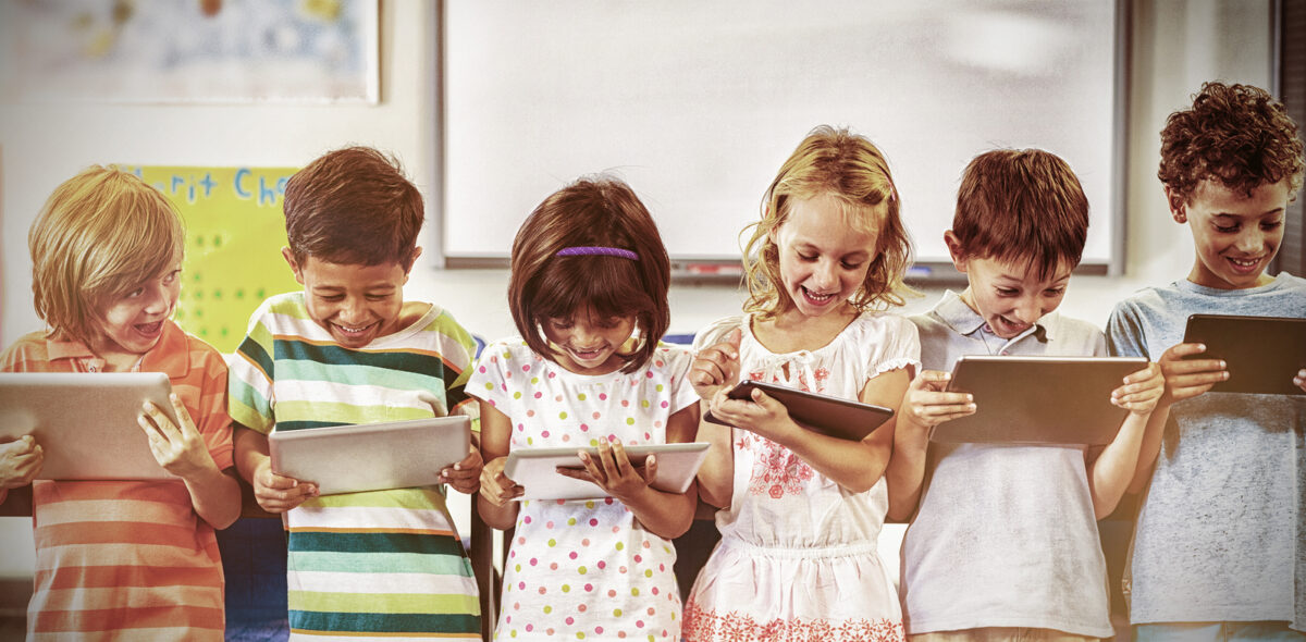 Six smiling primary school children standing in a row, each looking at their own tablet device.