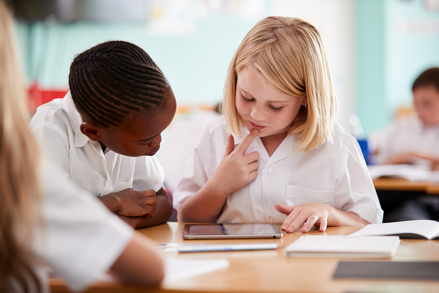 Two primary school pupils, one Black and one white, looking at a tablet on a desk in a classroom.