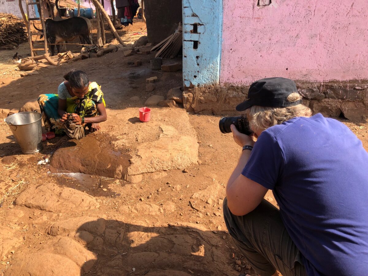 A person with a camera crouches, filming a woman washing clothes in a rural Indian village.