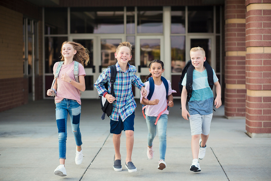 Four primary school children with backpacks running and smiling outside a school building.
