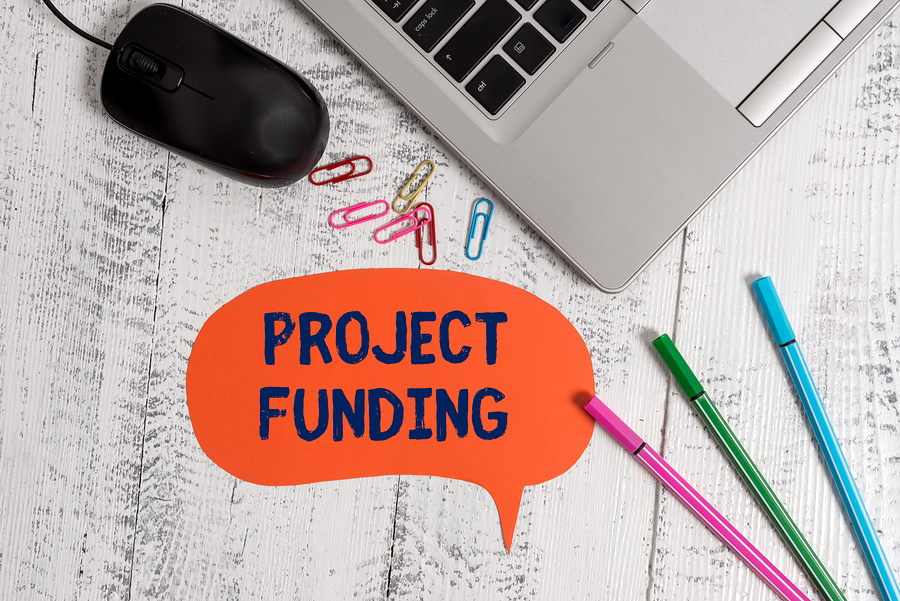 An orange speech bubble with the words PROJECT FUNDING in blue, next to a laptop, mouse, and pens