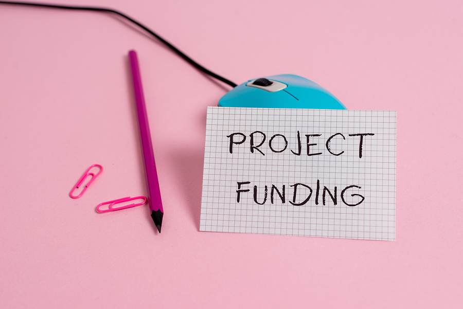 A blue computer mouse, a pink pencil, and two pink paperclips on a pink surface. A note attached to the mouse reads PROJECT