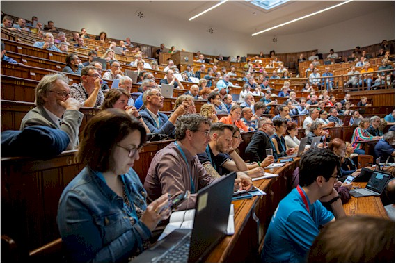 A large audience of people sitting in a tiered lecture hall, many using laptops or phones.