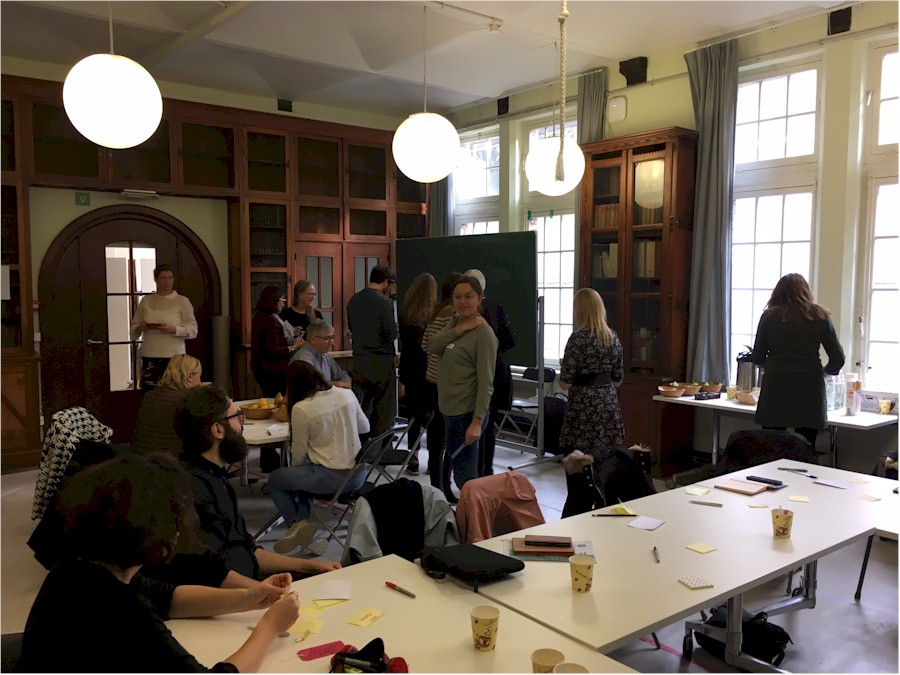 People gathered in a room, some standing and talking, others seated at tables with notes and cups.
