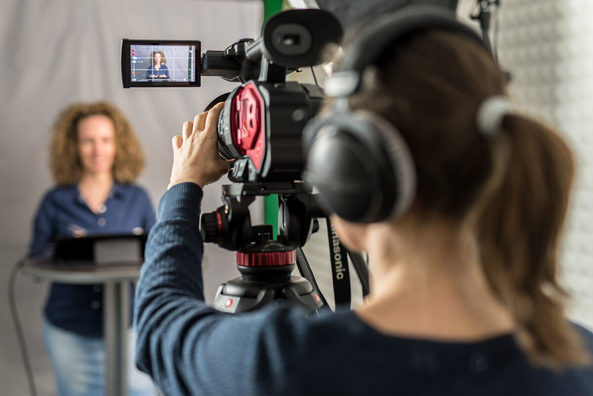 A person wearing headphones operates a video camera, filming a woman standing at a podium.