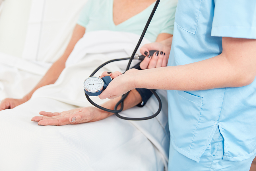 A nurse in light blue scrubs takes the blood pressure of a patient lying in a hospital bed.