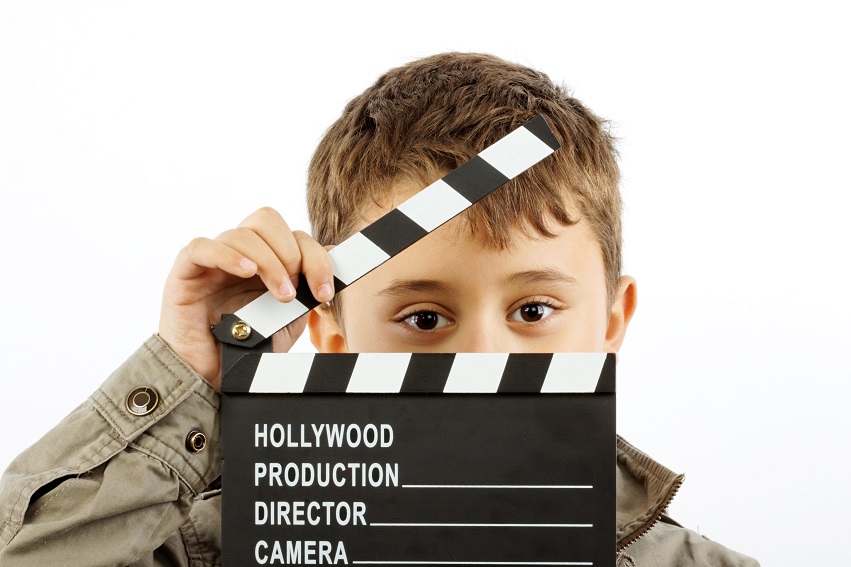 A young boy holds a movie clapperboard in front of his face, with only his eyes visible.