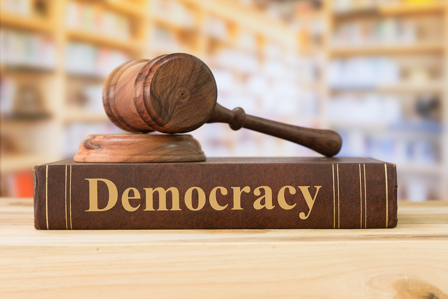 A wooden gavel rests on a brown book titled 'Democracy' with a blurred library in the background.