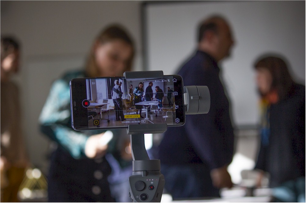 A smartphone on a gimbal records a group of people in a workshop setting, seen on its screen.