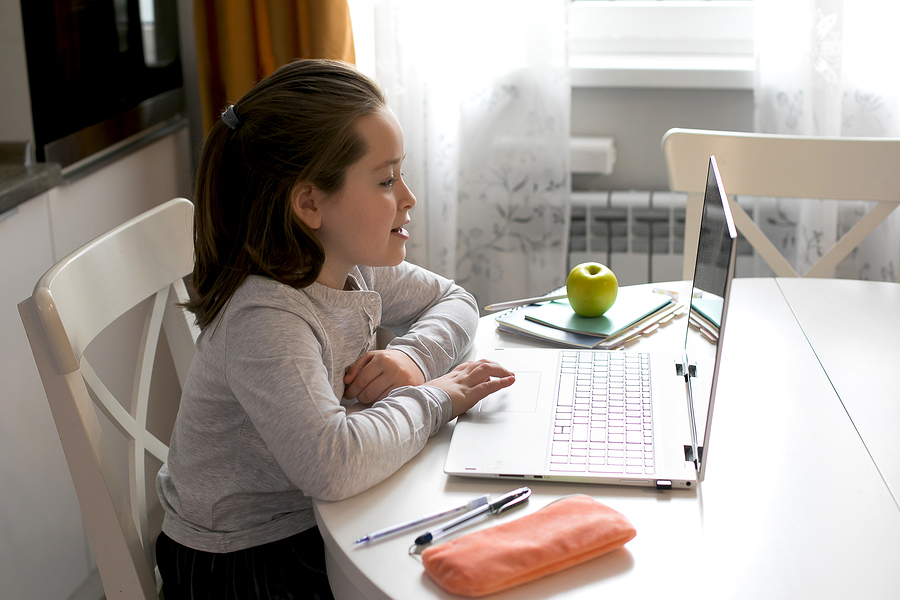 A young girl with a ponytail sits at a white table, looking at a laptop. An apple and books are nearby.