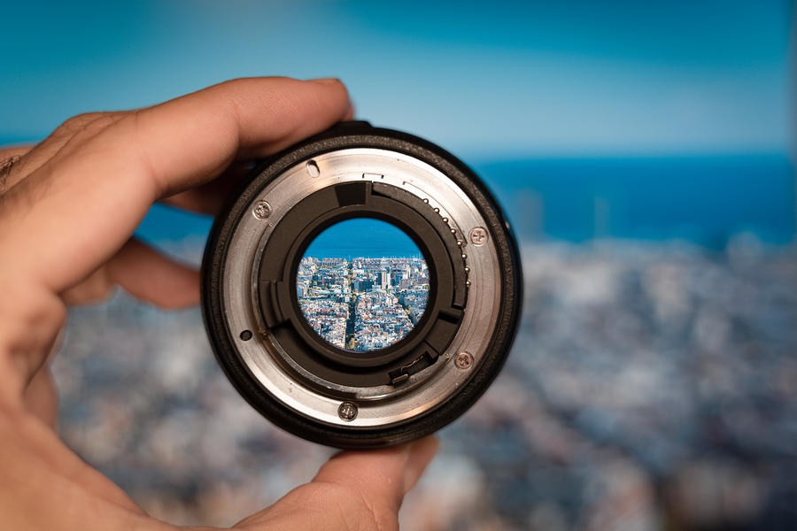 A hand holds a camera lens, through which a focused view of a city and the sea is visible.