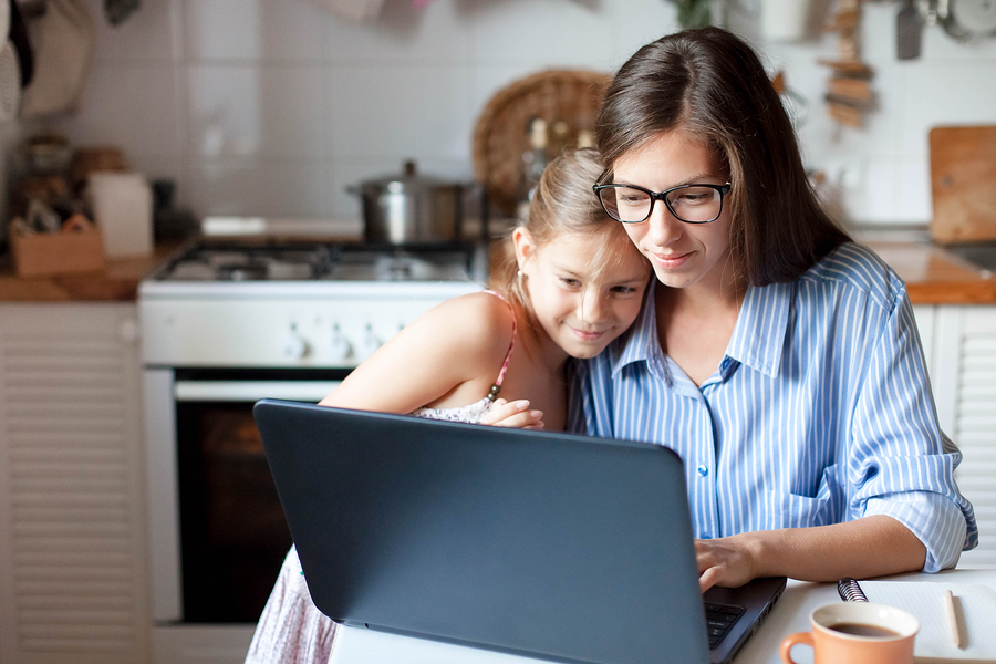 Mother and daughter looking at a laptop screen together in a kitchen, with a cup of tea nearby.