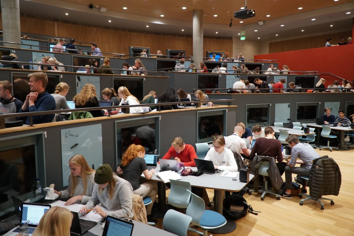 Students working on laptops and papers in a large, modern lecture hall with tiered seating.