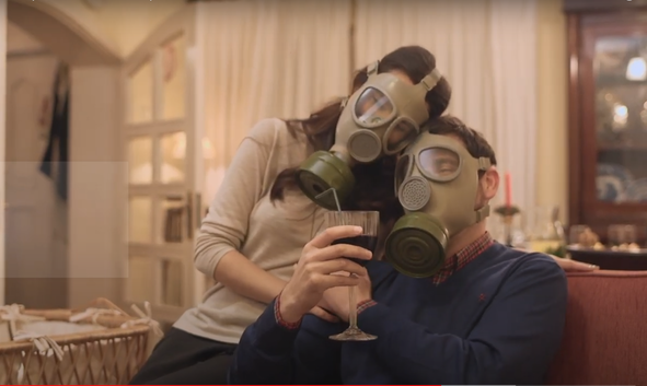 A couple wearing gas masks, sitting on a sofa, with the man holding a glass of red wine.