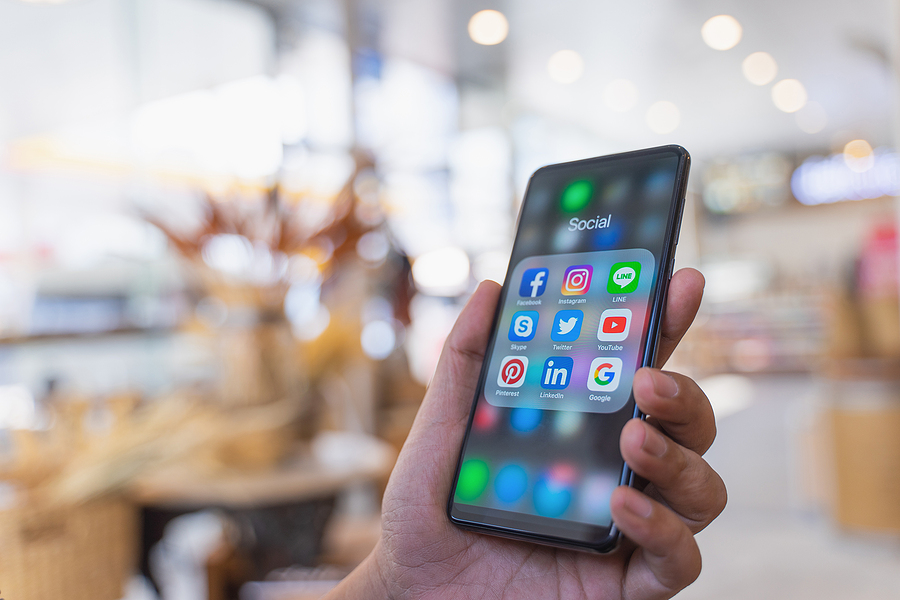 A hand holds a smartphone displaying a folder of social media apps, including Facebook, Instagram, and YouTube.