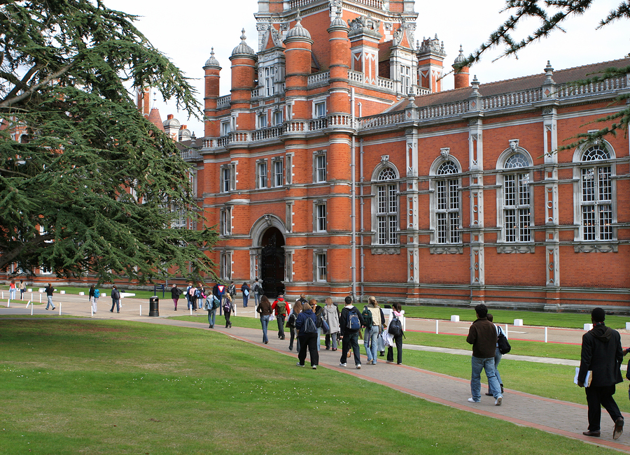 Students walk along a path on a green lawn in front of a large, ornate red brick university building.
