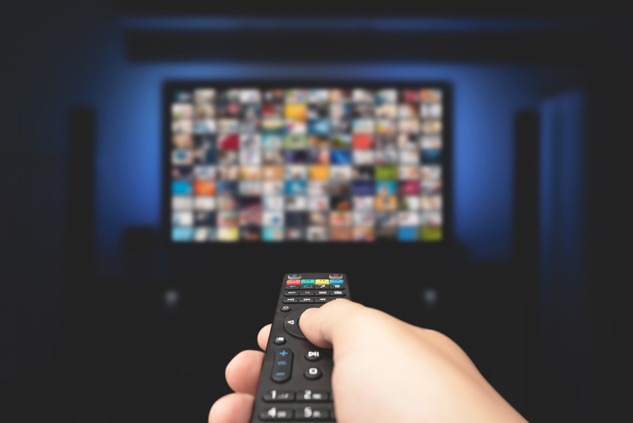 A hand holding a TV remote control, pointing at a large screen displaying a grid of blurred video thumbnails.