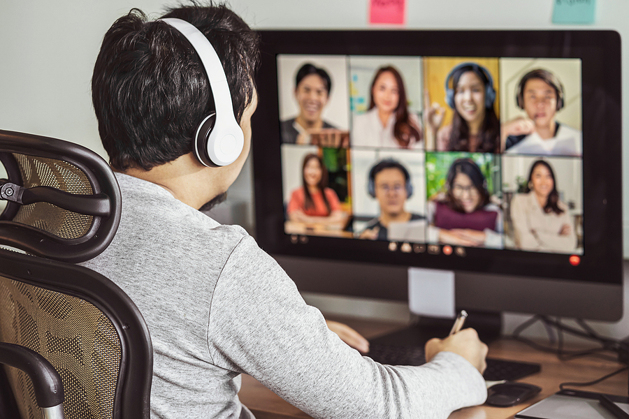 Rear view of a man wearing headphones, participating in an online video conference with eight other people.