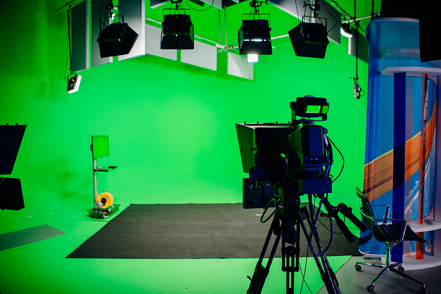A camera on a tripod in a green screen studio with overhead lights and a blue and orange set piece.