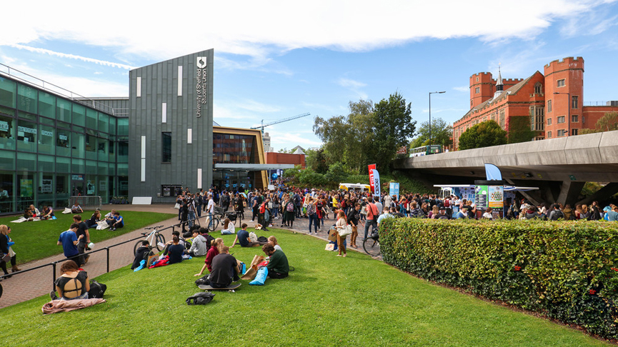 Students gathered on a university campus, with a modern building on the left and an older brick building in the background.