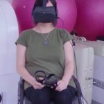 A person in a wheelchair wearing a VR headset and holding controllers, with pink exercise balls behind them.
