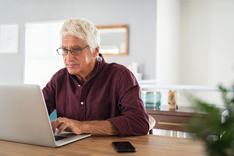 Older man with glasses and a maroon shirt typing on a laptop at a wooden table.