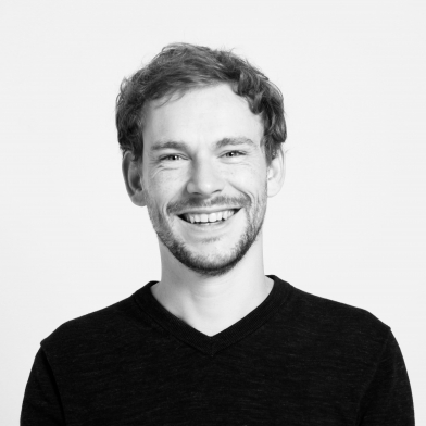 A man with a beard and curly hair smiles at the camera, wearing a dark V-neck shirt.