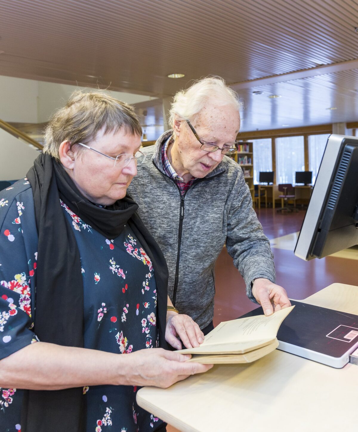 Two older people, a woman and a man, looking at a book at a self-service machine in a library.