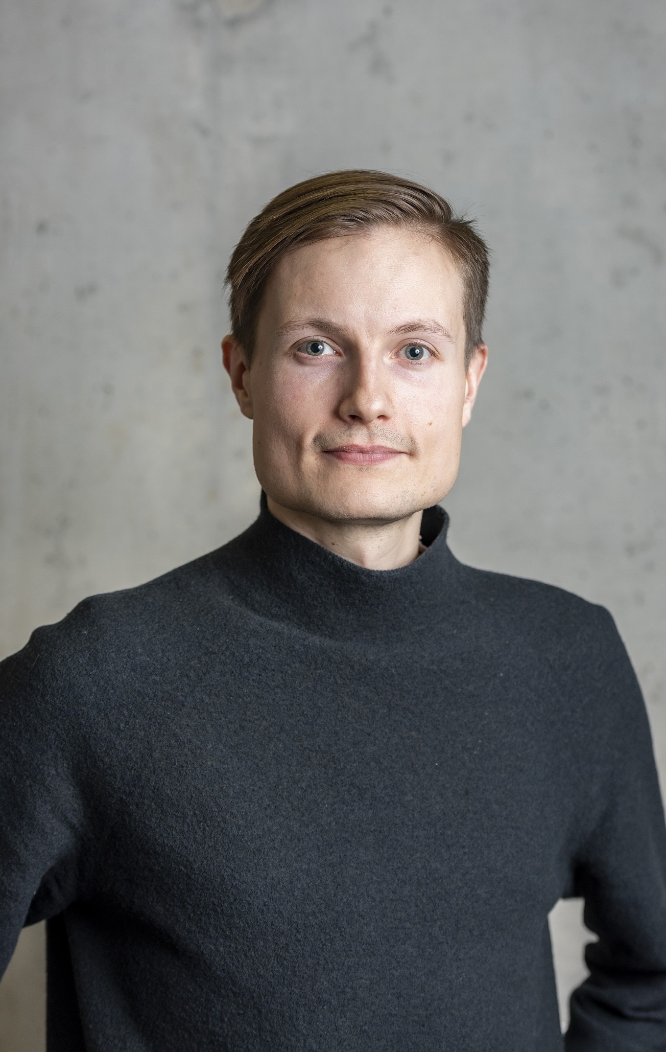 Headshot of Lauri, a man with light brown hair and a dark grey turtleneck jumper, smiling slightly