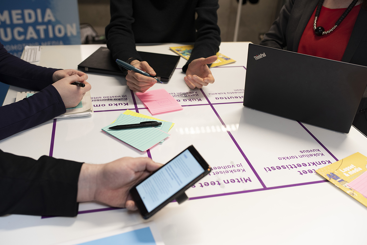 Four people at a table with a grid on it, writing on sticky notes and looking at a phone and laptop.