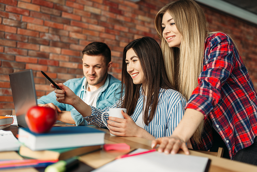 Three students, two women and one man, looking at a laptop screen, with books and an apple on the table.