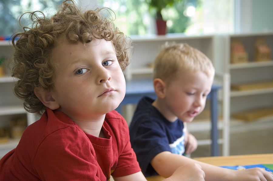 Two young boys in a classroom, one with curly hair looking at the camera, the other looking down.